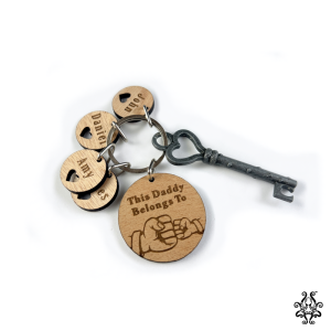 A wooden keychain set with an antique-style metal key and engraved wooden tags. The large circular tag reads "This Daddy Belongs To" with a fist bump illustration, and three smaller tags have names with heart cut outs.