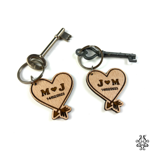 Two wooden heart-shaped keychains with engraved initials and date, attached to vintage-style keys. The keychains feature the initials "M ♥ J" and "J ♥ M" with the date "14/02/2023," along with an arrow through the heart design
