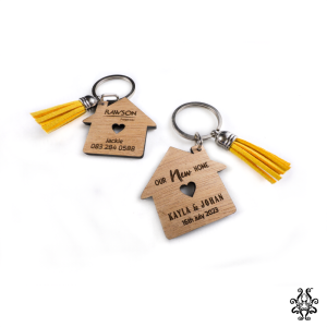 Two wooden house-shaped keychains with yellow tassels, featuring personalised engravings with real estate agent details and new homeowner information.