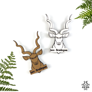 Two wooden wedding kudu lapel pins, one in natural wood and the other in white, engraved with "Jaco - Bruidegom," displayed on a white background with green ferns.